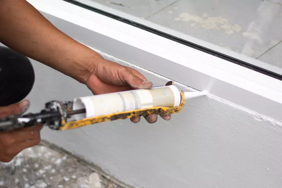 A person is sealing a white exterior window frame with a caulking gun