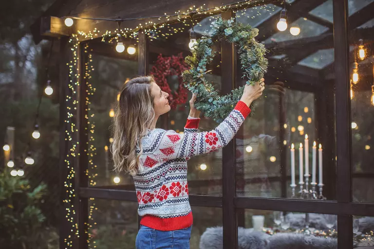 A woman putting up a holiday wreath in front of a glass sunroom. 