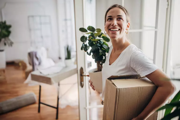 Woman carrying a box and a potted plant into a home.