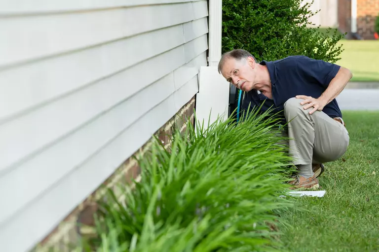 An older man investigating the foundation of his home to look for water damage that could be a result of clogged gutters and roof damage.