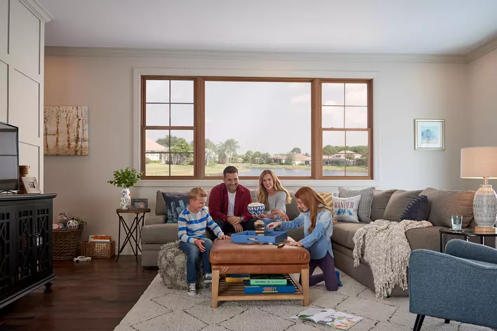 A family playing games in their modern, decorated, naturally lit living room that contains a large picture window flanked on each side by a double-hung window and overlooking a pond surrounded by palm trees and grass.