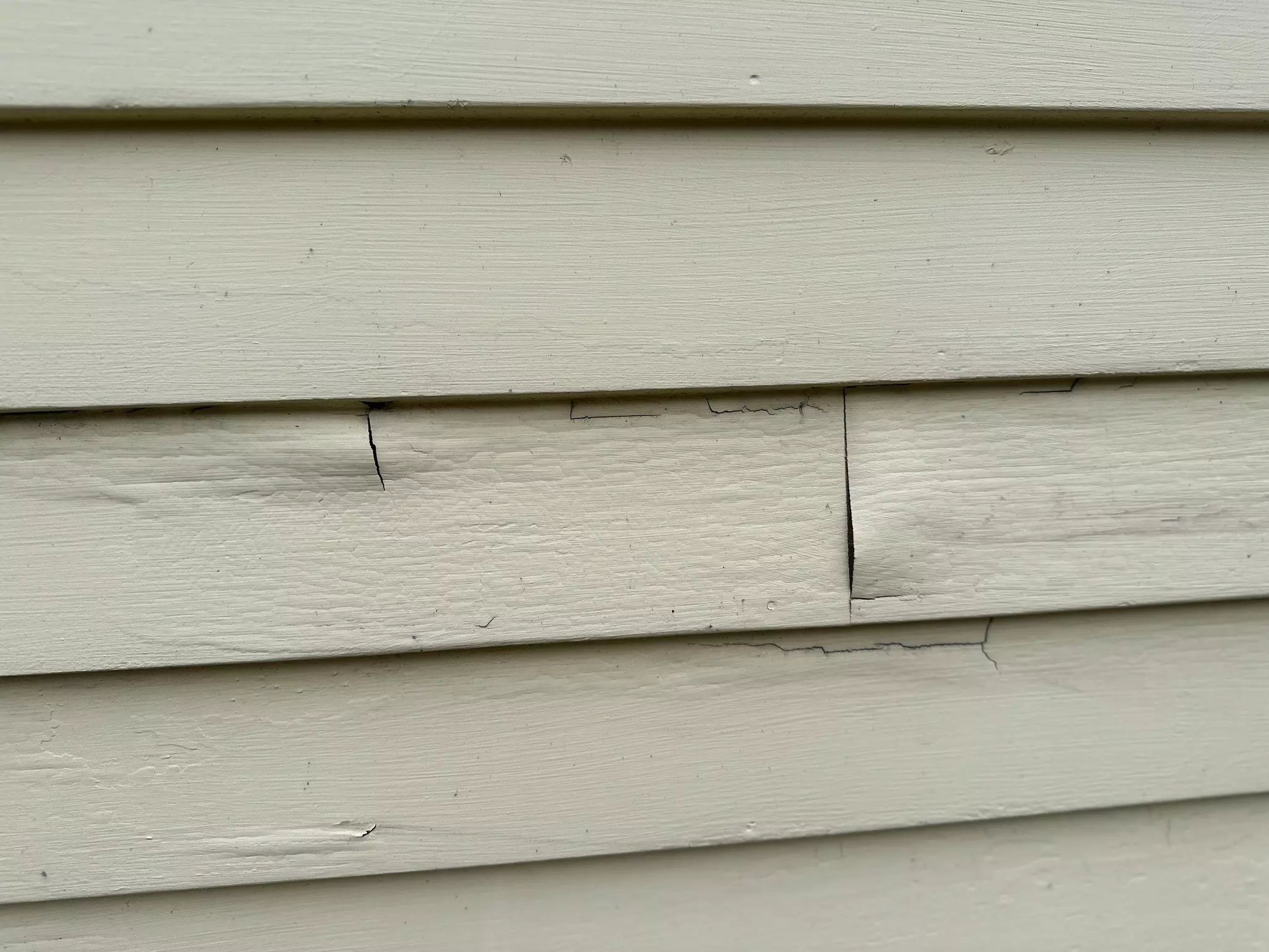 Damaged cream-colored siding showing visible cracks, warping, and a loose panel from age and weathering.