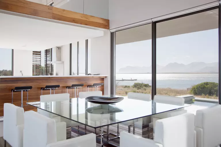 A dining room in a modern home with a glass table and white chairs. The back picture windows look out over a lake with nearby mountains.