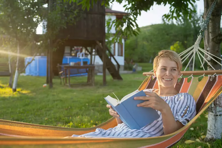 A woman lying in a hammock holds a book open and looks at someone out of frame. She appears to be enjoying her outdoor living space.