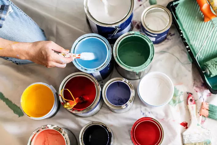  A person dipping their brush into a light blue paint can within a group of other multicolor paint cans on the floor