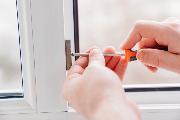 Two hands using a small screwdriver to remove the balance shoe from the bottom of a vinyl window frame.
