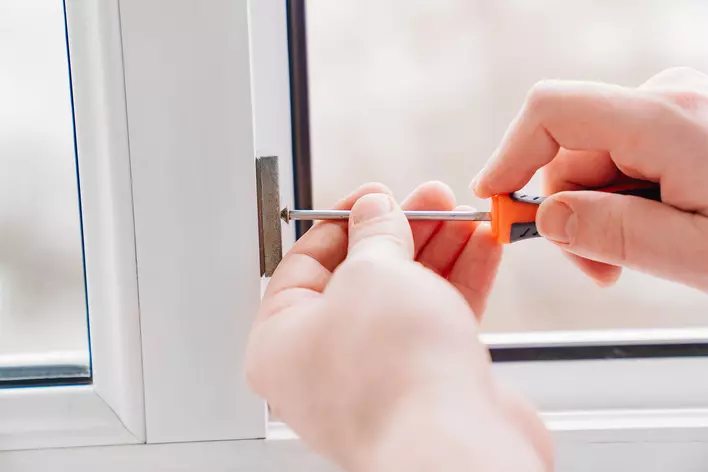 Two hands using a small screwdriver to remove the balance shoe from the bottom of a vinyl window frame.