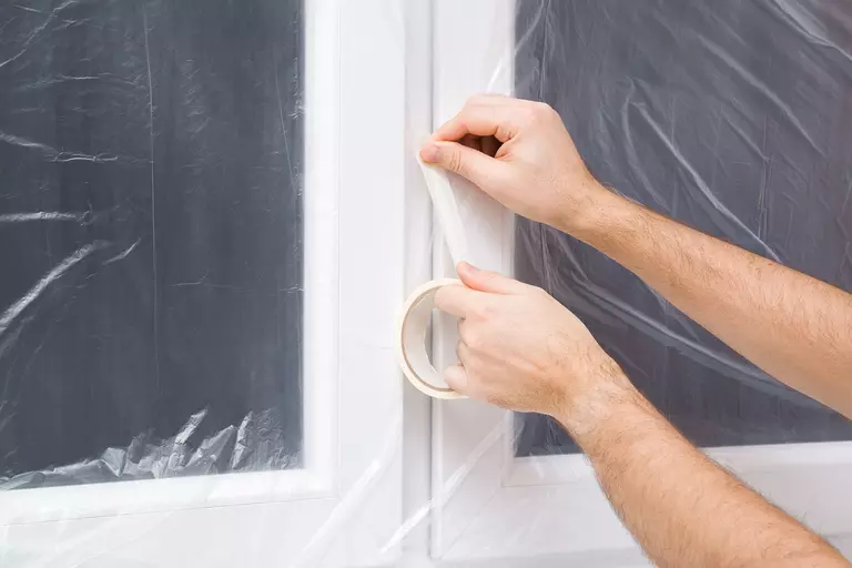 Person taping plastic film over a window in an effort to stop window drafts.