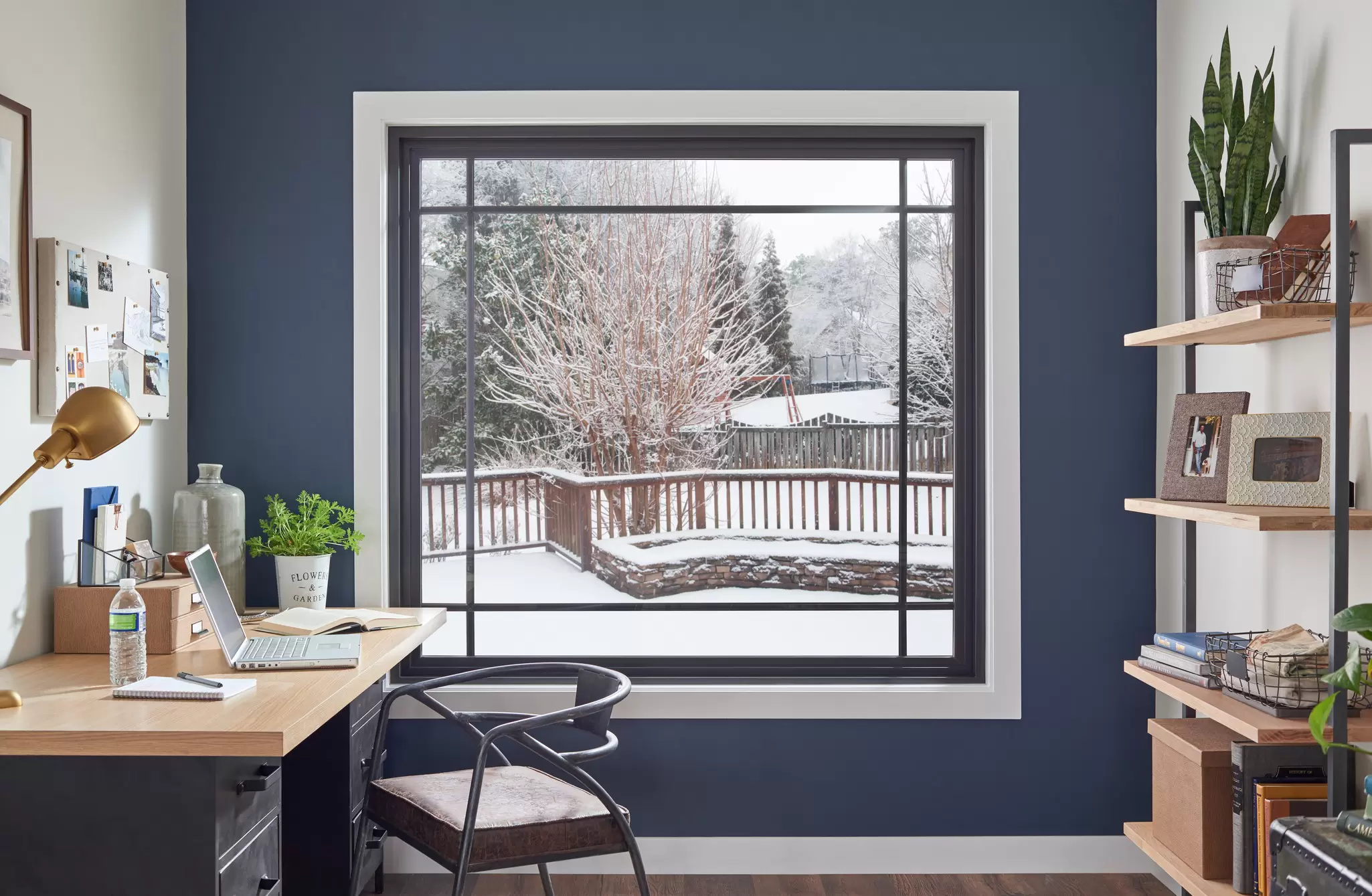 Home office with a black-framed picture window with grilles overlooking a snowy deck.