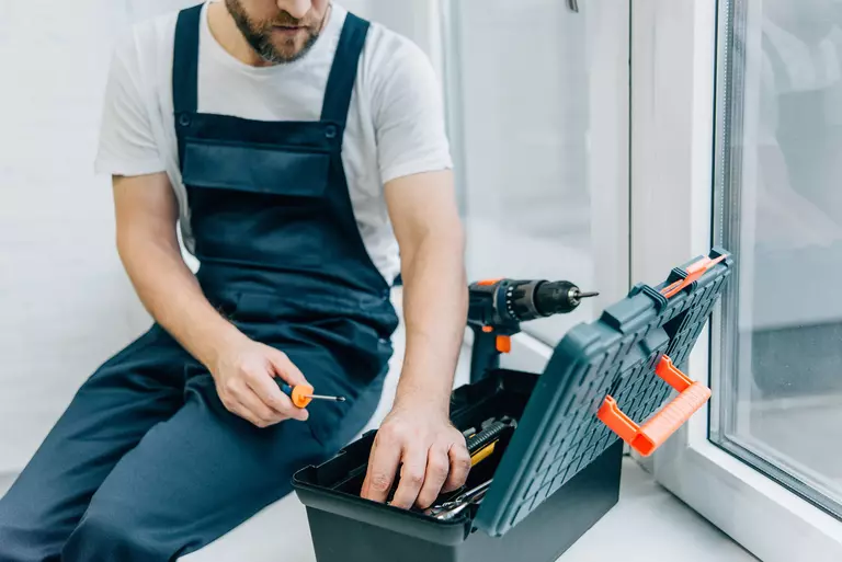 Worker sitting down near a clear window as he looks inside his toolbox next to a power drill