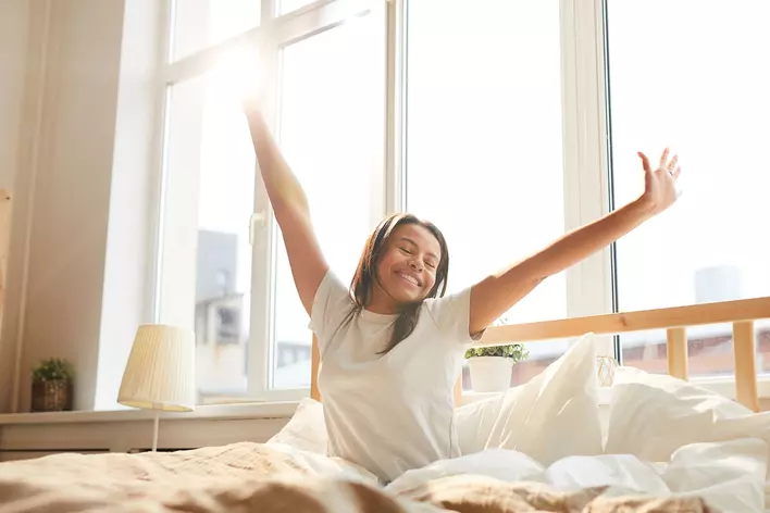 A woman in bed, stretching after getting better sleep thanks to the natural light coming through the windows behind her.