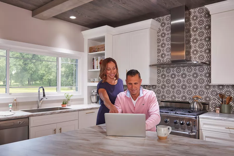 Picture of a man and a woman sitting in a large, remodeled kitchen, looking and pointing at an open computer screen. 