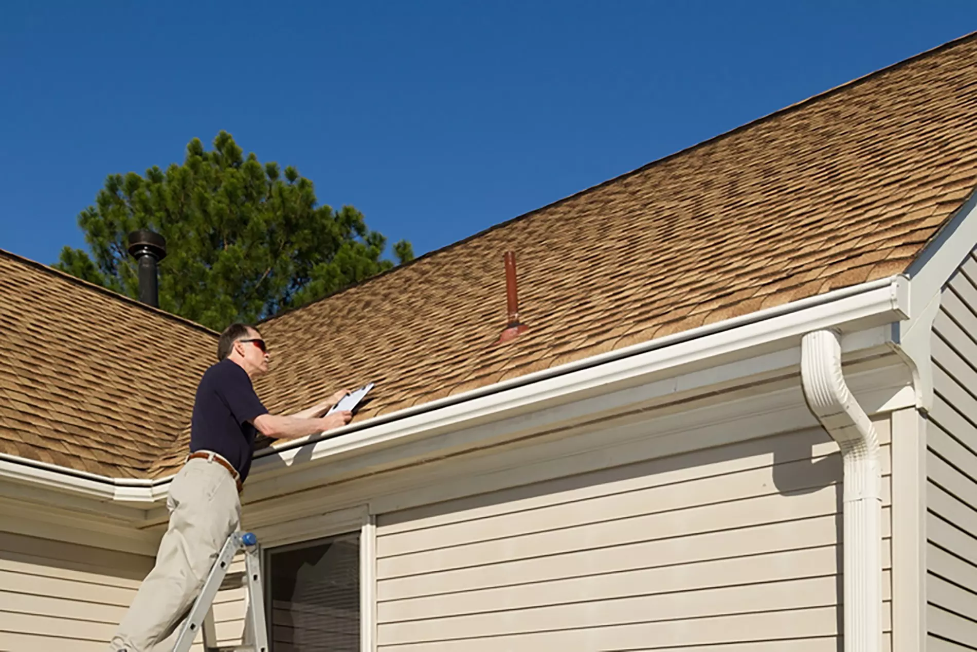 A contractor on a ladder inspects a roof maintenance checklist on a sunny, cloudless day.