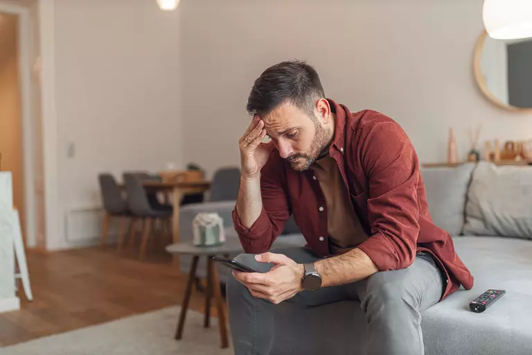 An upset man sitting on a couch looking at his phone.