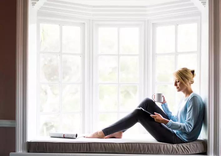 Woman holding coffee and digital tablet while sitting in front of a large bay window
