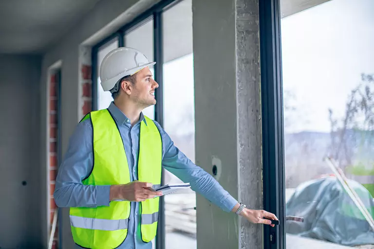 A smiling man with a hard hat looking through the glass of an exterior door.