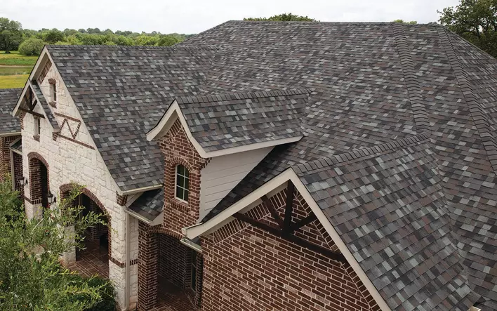 A very large home with mixed brick colors and wooden accents that has a new roof made of quality material and different shade shingles. 