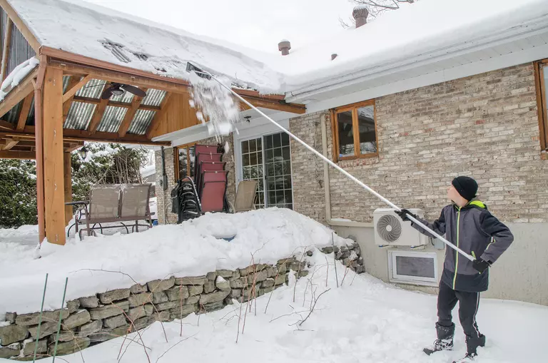 A man standing on the ground using a roof rake to remove snow from his back porch
roof.