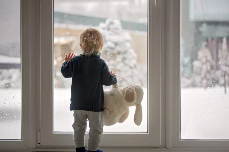 A child with a stuffed animal looking through a door at the snow.
