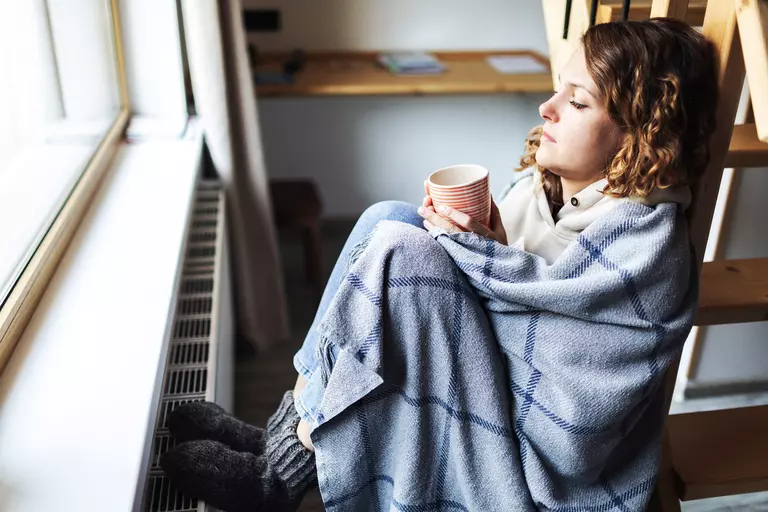 Woman with a beverage mug wrapped in a blanket and sitting by a window.