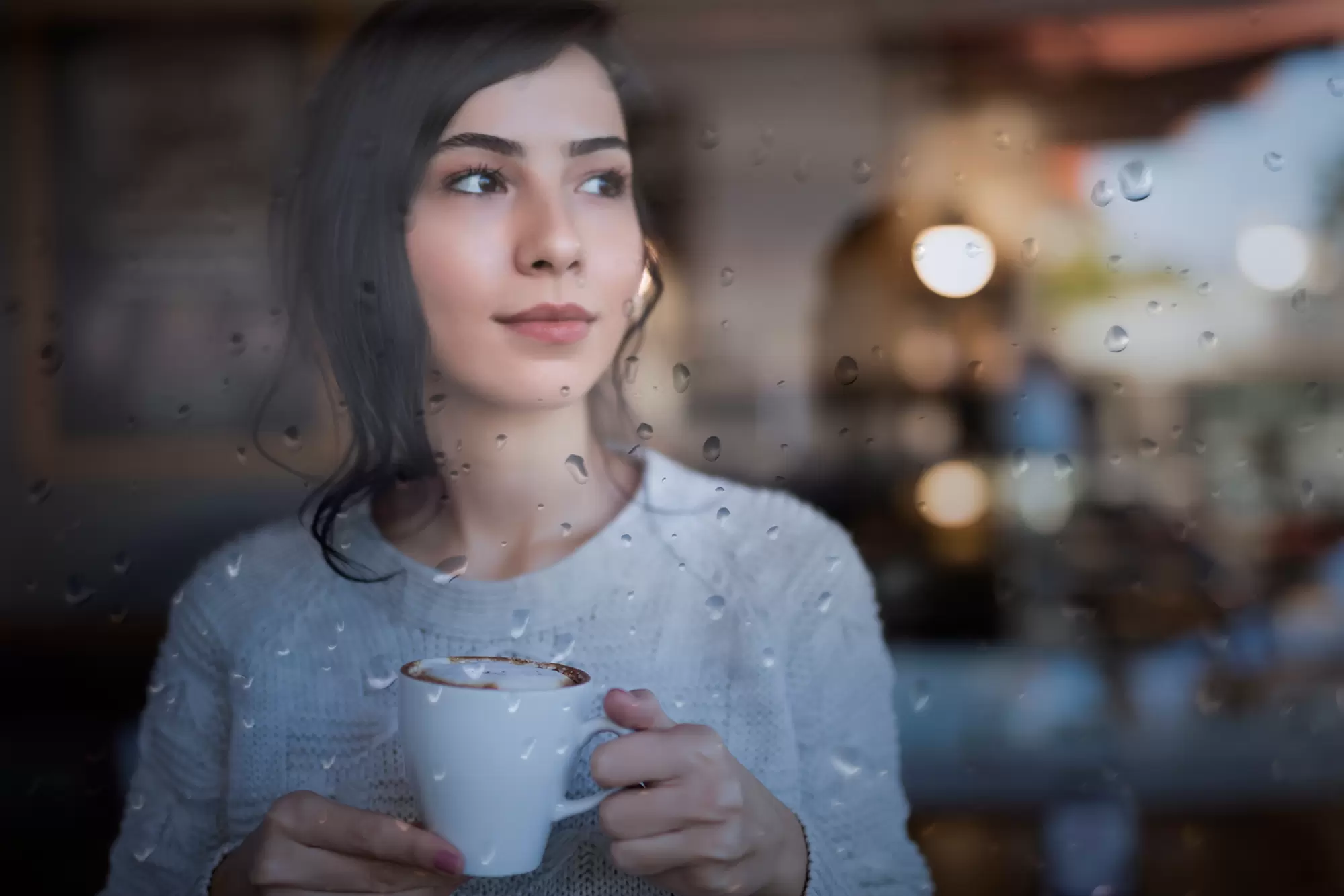 Woman holding a coffee mug on the other side of a window with raindrops.