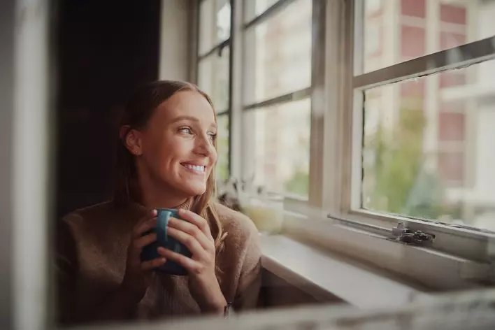 Young woman smiling while drinking coffee as she looks outside windows of her home.