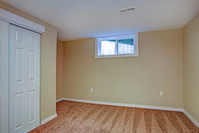 An empty room in the basement of a home that is naturally lit by a sliding basement window.