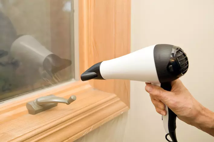 A person uses a hairdryer to adhere heat-shrink plastic film to their windows to prevent
            condensation from building up.