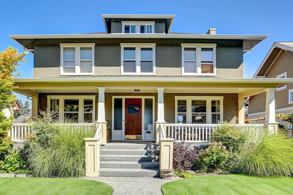 A craftsman-style home with a red entry door, a large front porch, and a landscaped front yard with flowers and bushes surrounding the walkway.&nbsp;