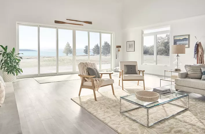 A very bright and naturally lit, light-toned living room that has an entire wall of sliding patio doors and large windows that overlooks beautiful scenery beside a lake.