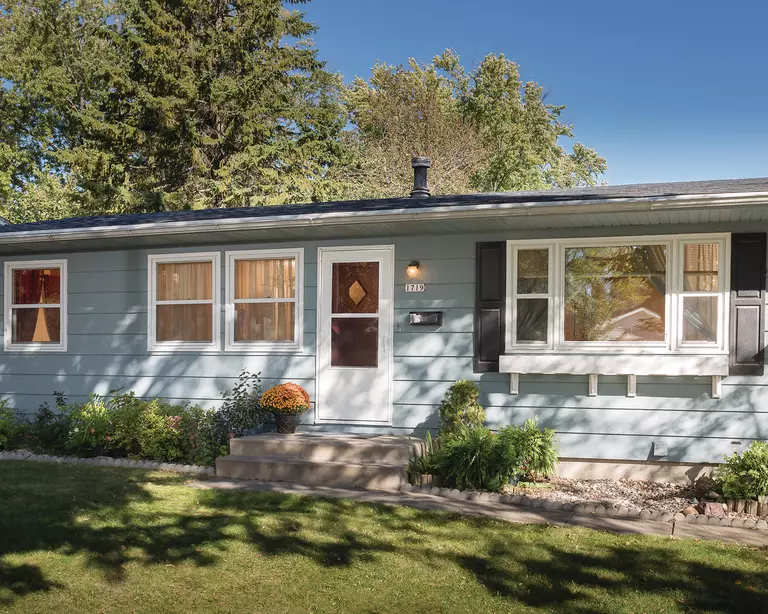 A home with powder-blue siding, dark shutters, and white trim, including a storm door and window boxes to protect and beautify the house.