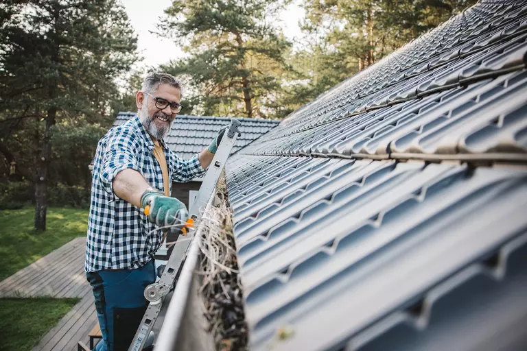 A smiling man on a ladder prepares to clean gutters with a wand tool.