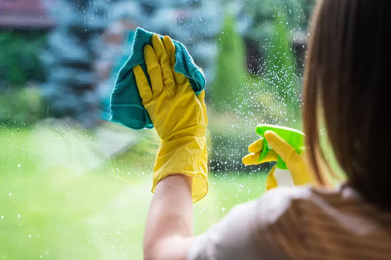 A woman wearing disposable gloves while using a cloth and spray bottle to wipe glass.