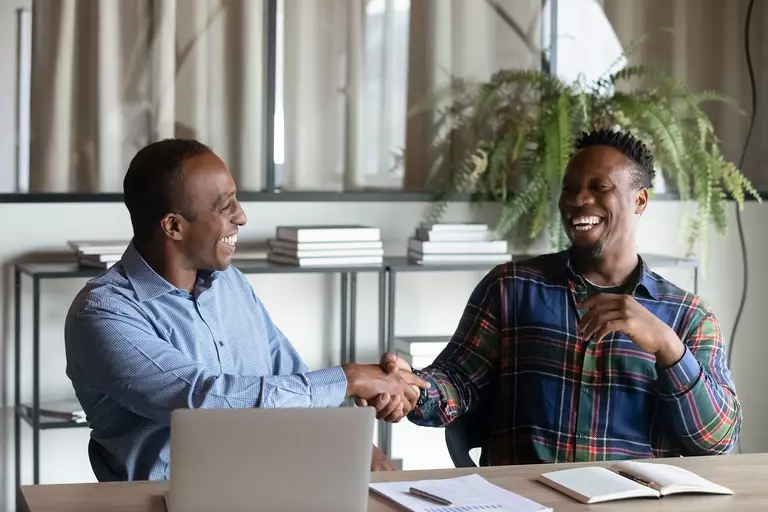 Two men happily shaking hands with each other while sitting at a desk. 