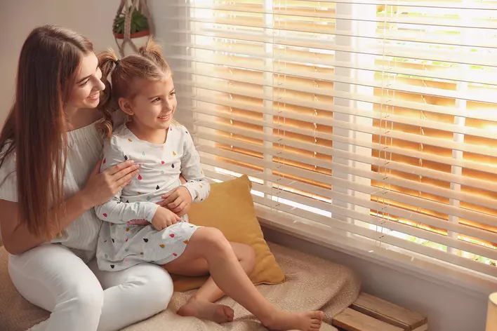 A woman and child sitting next to a window sill shaded with Venetian blinds