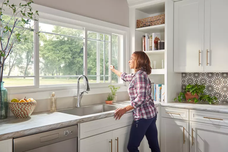 A woman opening the window in a kitchen that is behind the sink.