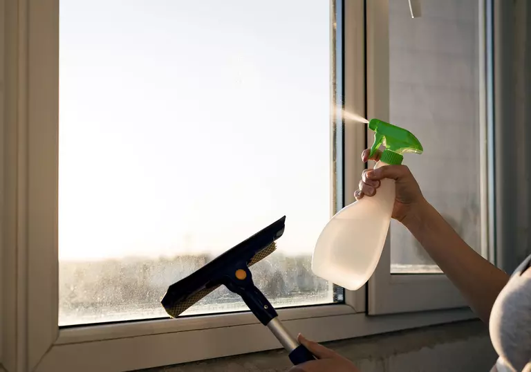 A person holding a squeegee and spray bottle applies a cleaning solution to a window.