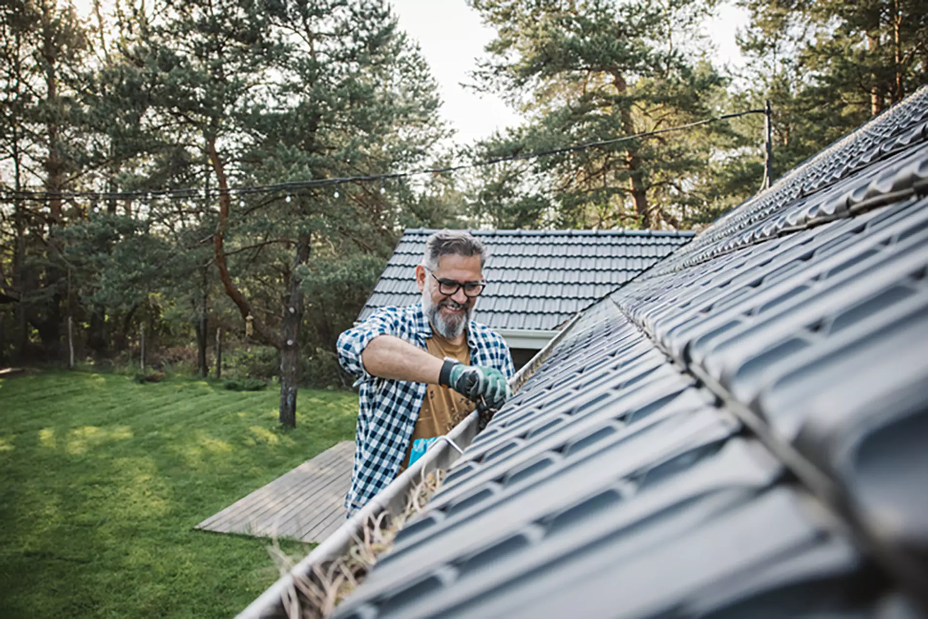 A homeowner on a ladder grimaces while pulling debris from his gutters, highlighting the importance of diligent roof maintenance. 