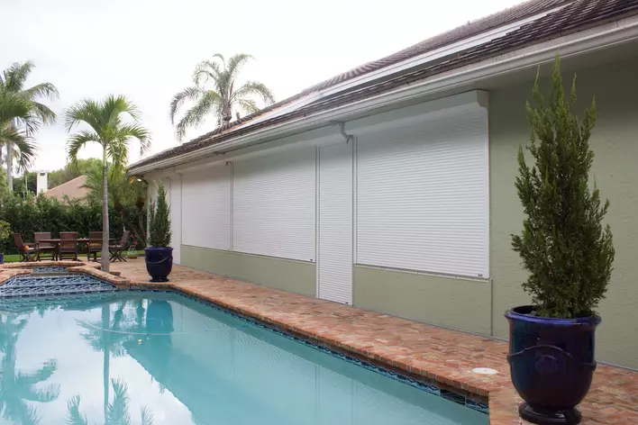 A home with a blue pool, palm trees, and roll-down shutters.