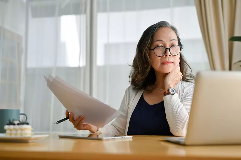 Woman holding documents and viewing a laptop.