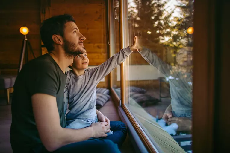 A man and young boy look out the picture window of their log cabin at woods outside.
