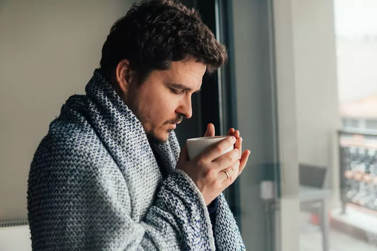 Man standing with a blanket around his shoulders and holding a beverage mug.