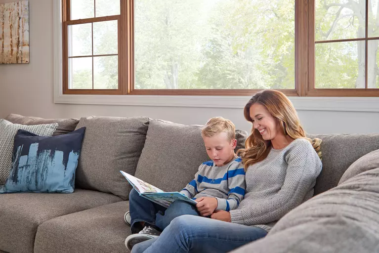 Mom and child reading while sitting on a gray couch inside a home underneath a large picture window.