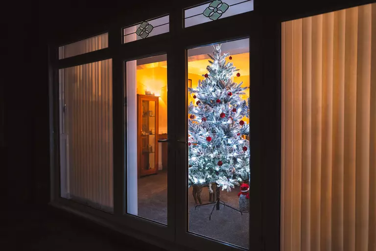 A Christmas tree viewed outside a home at night through casement windows.