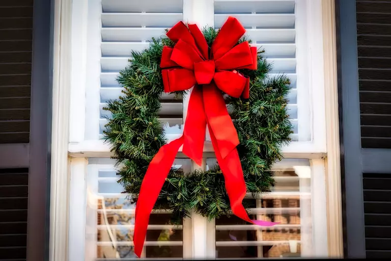A holiday wreath with a large red ribbon in front of a double-hung window.