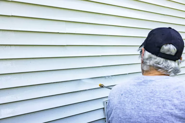 Man using a pressure washer to carefully clean white vinyl siding.