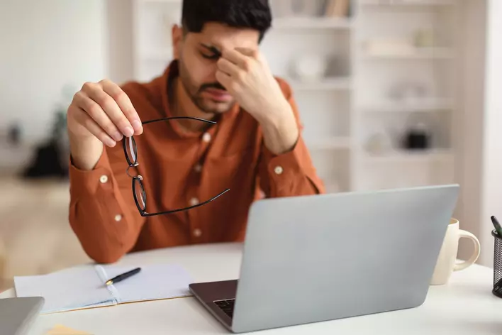 A man holding his glasses and pinching his nose to alleviate eyestrain caused by the brightness of his monitor. More natural light in his workspace should also help alleviate this concern.