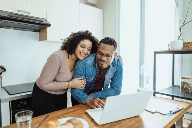 Couple at home using a laptop at a kitchen counter with bagels and a water cup shown.