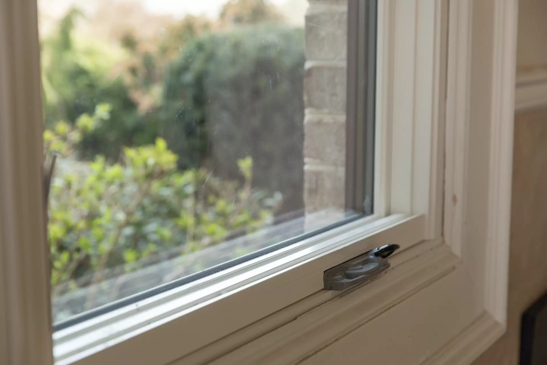 Close-up of a white double-hung window sash lock with greenery visible outside.