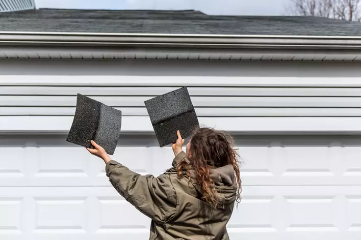 A woman holding up two types of shingles in front of her home to determine what type of roof will look best. 